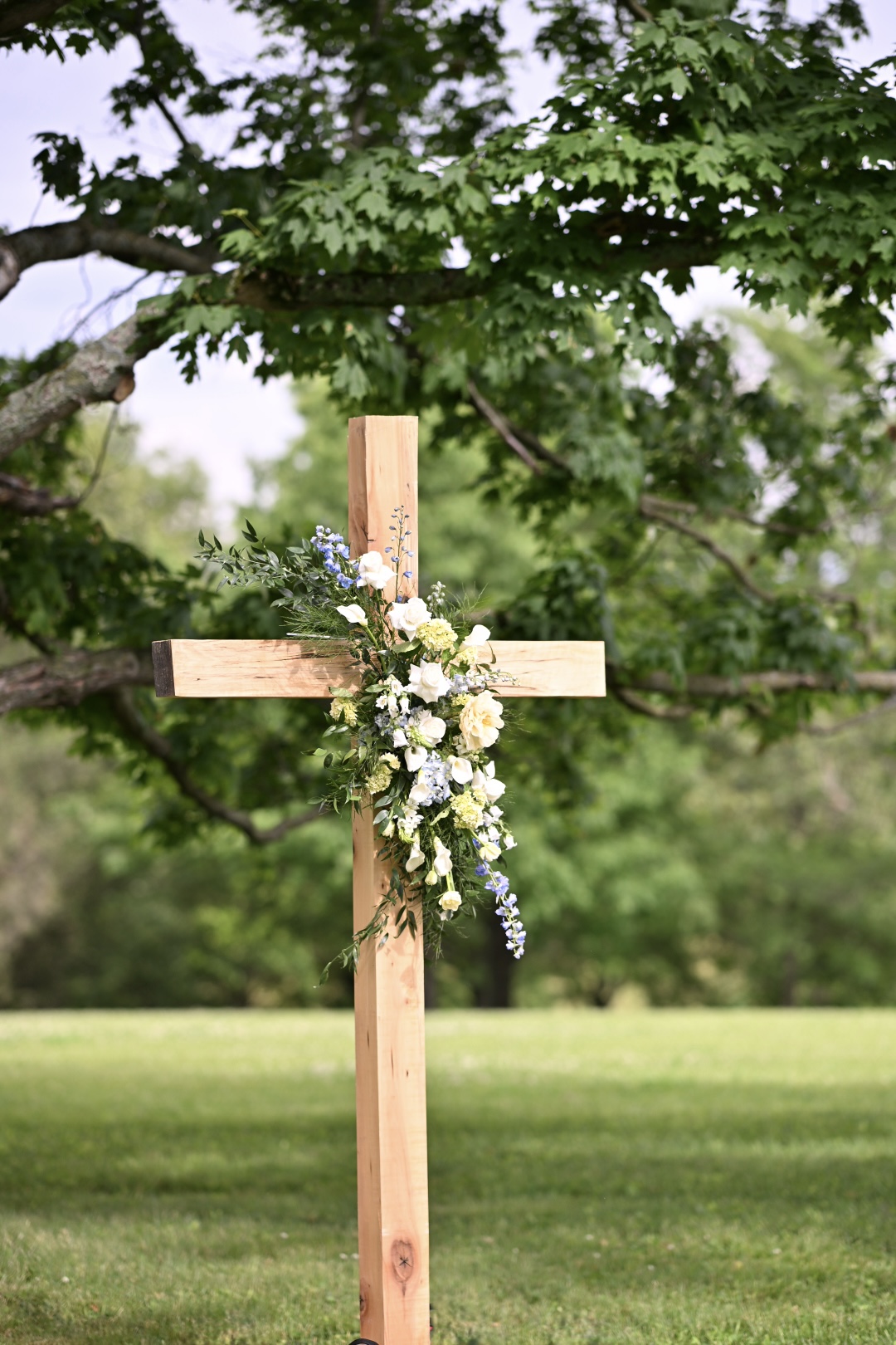 Wooden cross adorned with white and blue florals beneath a maple tree